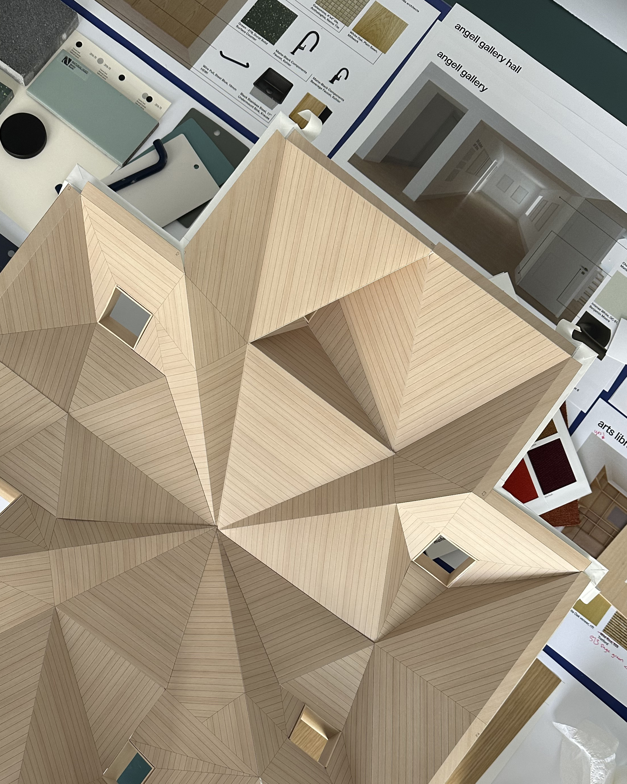 Close-up photograph of a light-colored wooden architectural model. The model's roof is a complex geometric design with triangular and diamond shapes. Material samples and printed design plans are scattered around the model.