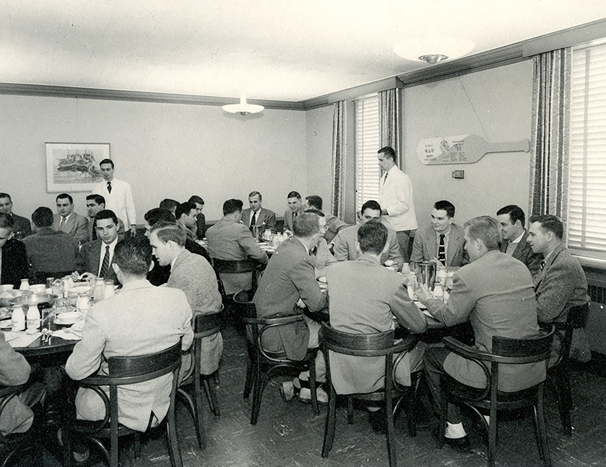 Vintage black and white photograph of a group of men seated around a long dining table in a room with blinds.
