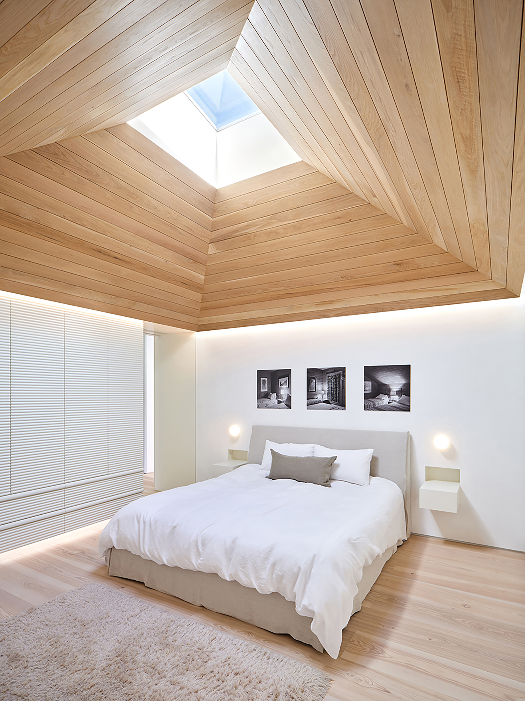 Bedroom in neutral, natural and white tones, with angular, wood-clad skylight above, and floating wall-mounted side tables and sconces on either side of the bed