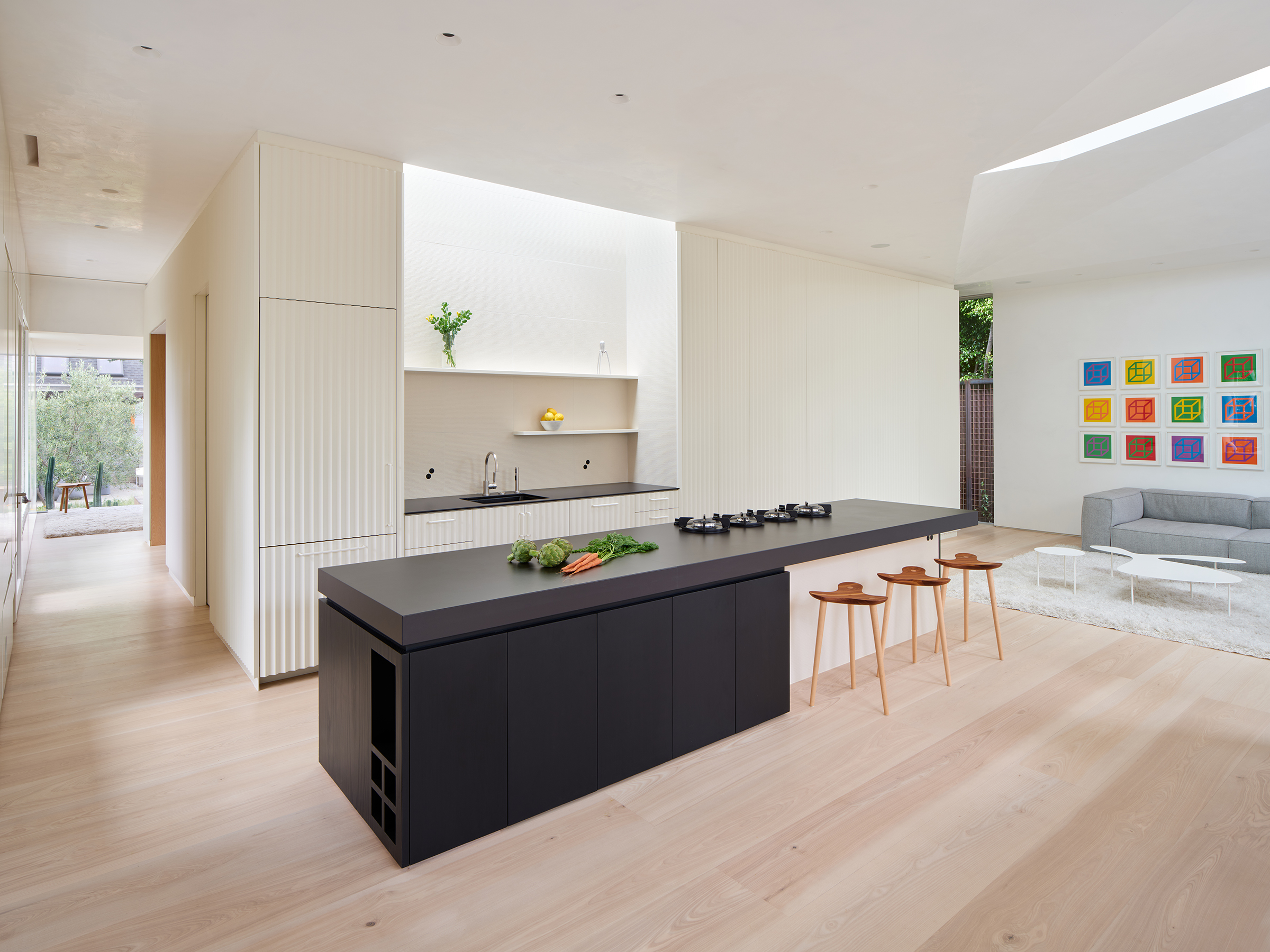 View of the kitchen island at the center, kitchen counter behind it with concealed skylight above, hallway leading to exterior to the right, and living area with sofa to the left