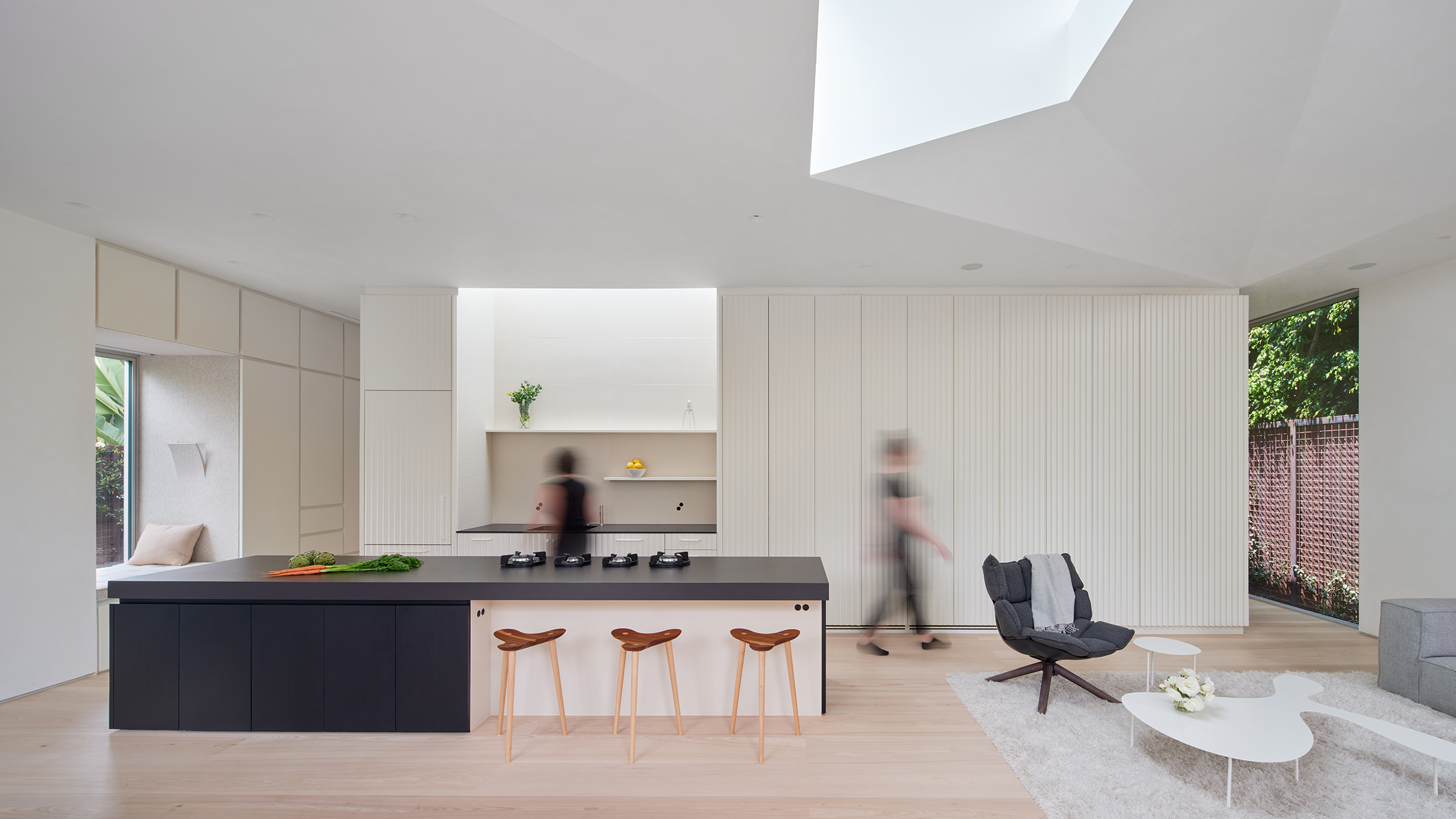 Elevational view of the main living space with window reading nook to the left, kitchen island and kitchen counter at the center, faceted skylight above, and storage millwork adjacent to the floor to ceiling window to the right.