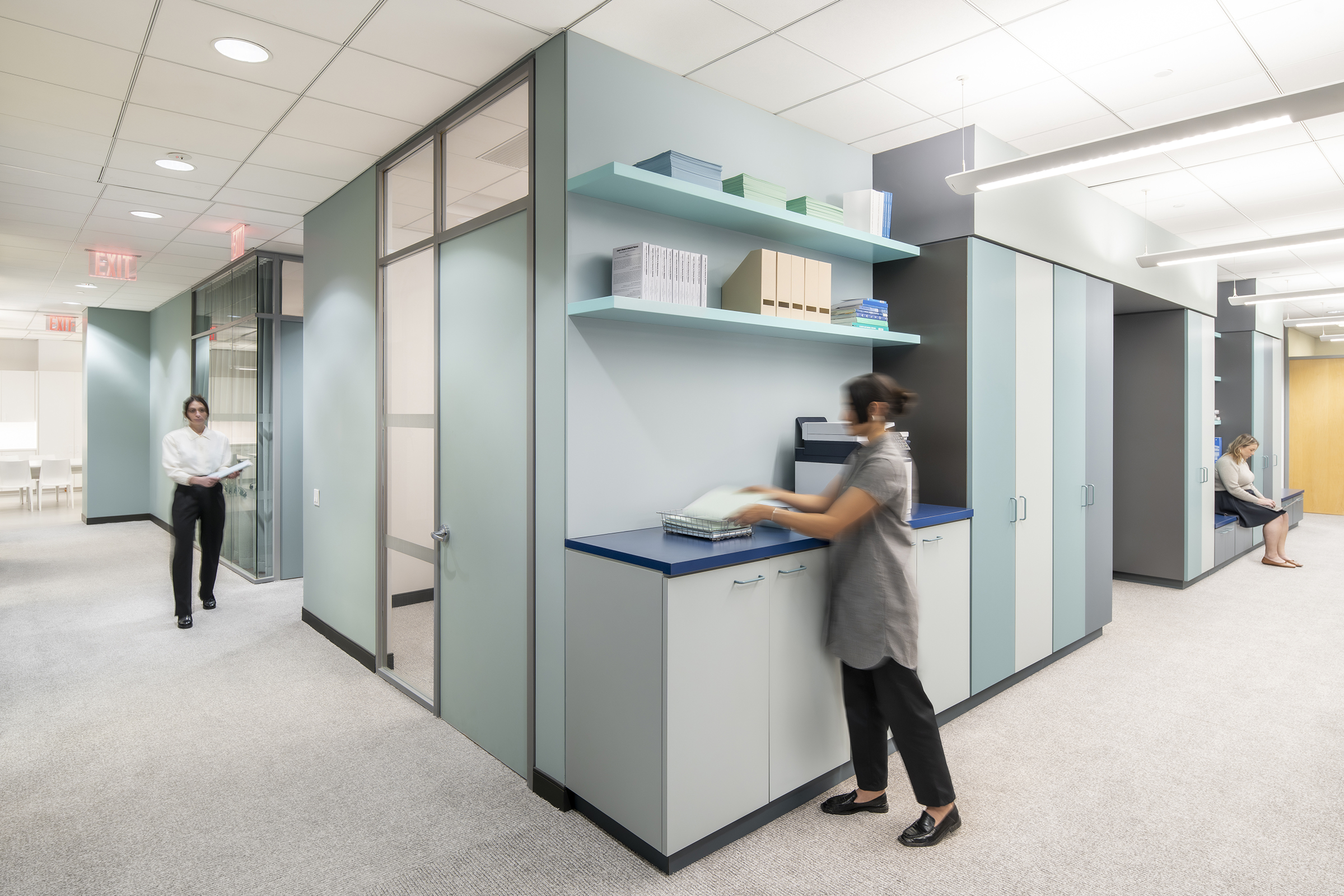 A view of the central millwork cube. In the foreground, a woman in a gray tunic and black pants is placing papers into a wire basket on a counter with cabinets below and shelves above holding books, files, and office supplies. To the left, a man in a white shirt and black pants walks down the hallway holding a folder. In the background, a woman in a light-colored top and dark skirt sits on a built-in bench, working on her laptop. The space has a neutral color palette with soft blue and gray tones, glass partitions, and bright overhead lighting.
