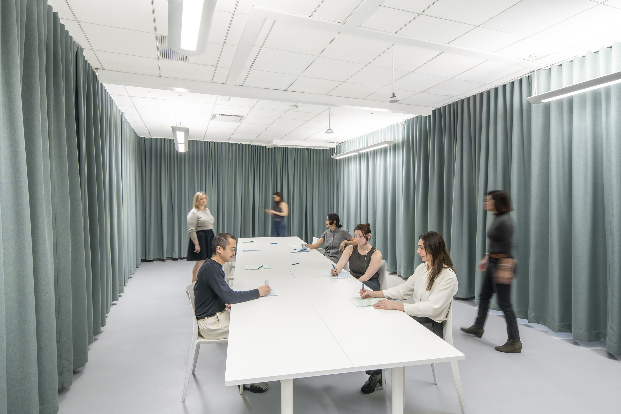 A collaborative workspace with employees engaged in a meeting or group discussion. The room is enclosed by floor-to-ceiling green curtains, creating a private setting. Tables have been reconfigured as a long central work surface with scattered notepads and pens. Some employees are seated, actively writing or conversing, while others move around or observe from a distance. The lighting is bright and uniform, complementing the minimalistic and modern aesthetic. The scene suggests a flexible, open-ended work session, possibly a brainstorming or strategy meeting.