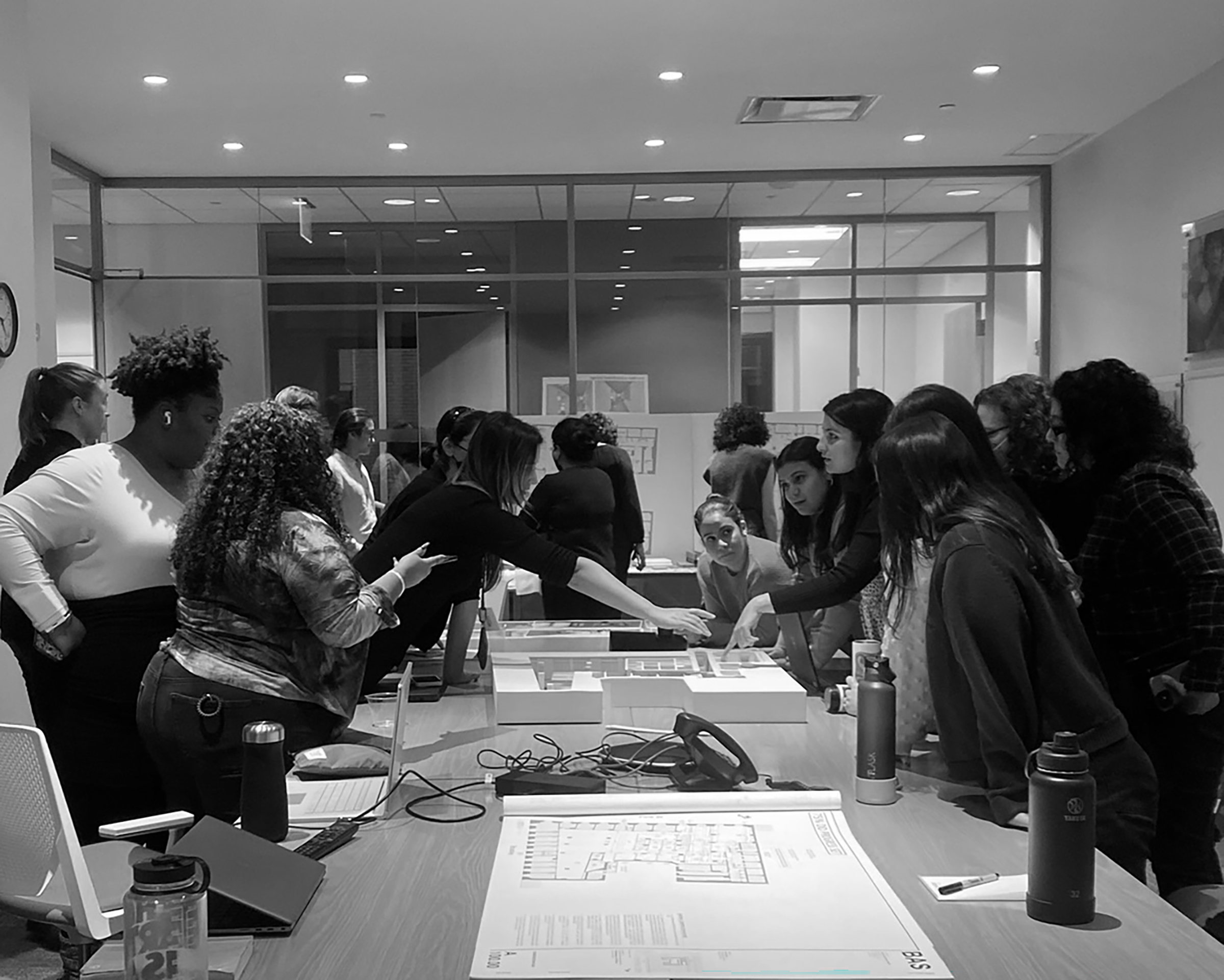 A black and white image showing the group of clients gathered around a table, discussing and pointing at architectural plans and models, during a design meeting. Floor plans are laid out in the foreground. The meeting is taking place within their existing glass enclosed conference room.