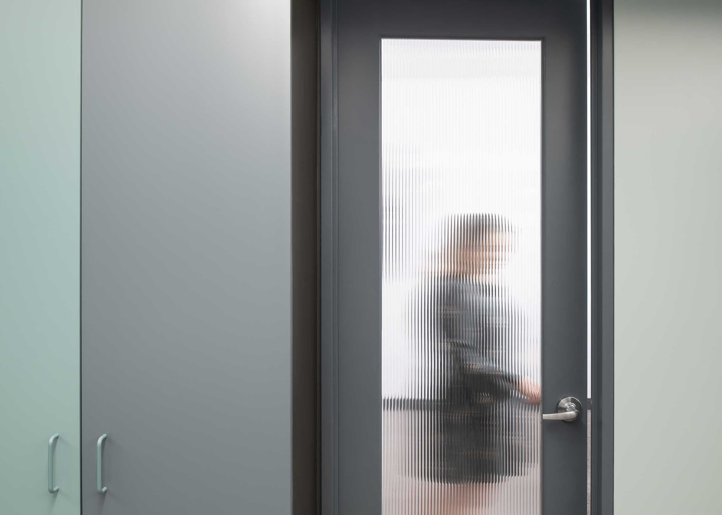 A view of a sillhouetted figure standing behind the reeded privacy glass of a meeting room door with colorful millwork doors adjacent to it