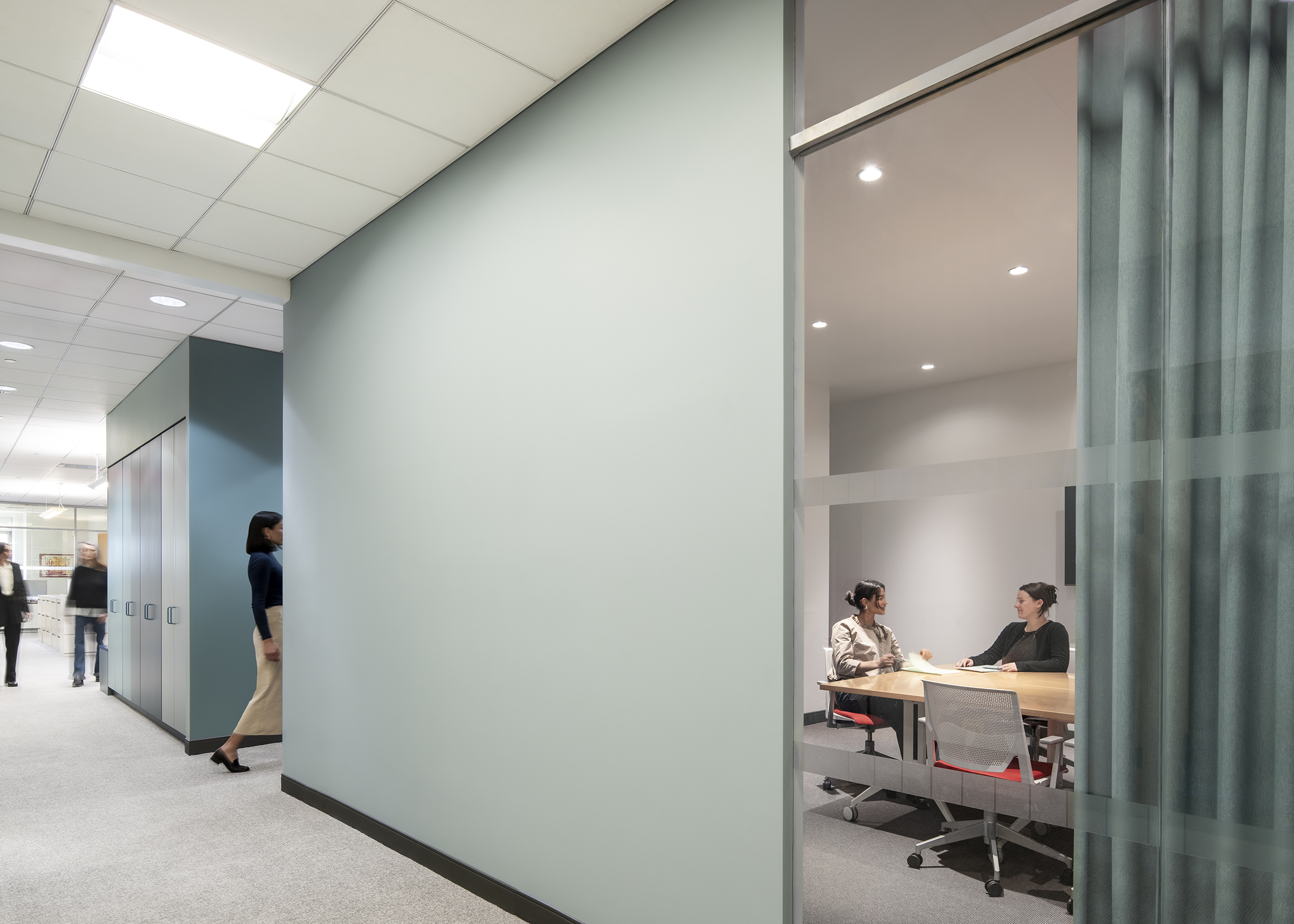 View of a meeting taking place within the meeting room, partially concealed by drapery and people circulating around the colorful millwork in the background