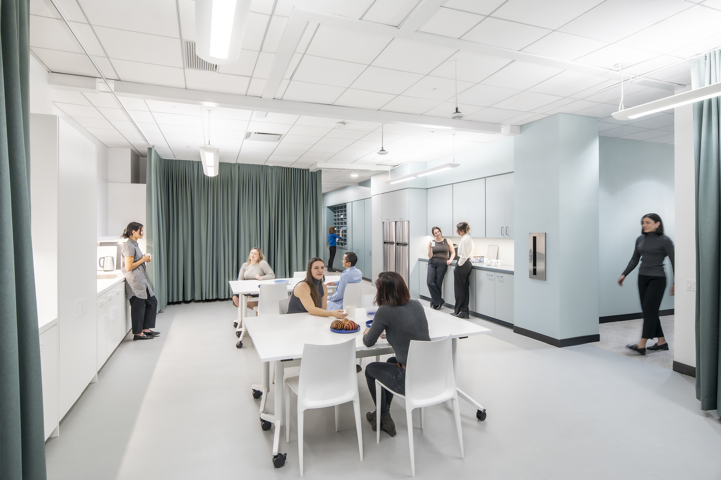 A modern office breakroom with employees engaging in casual conversations and taking a break. The space has white tables and chairs, a kitchenette with light blue cabinetry, and a refrigerator. In the foreground, two women sit at a table, one with a plate of croissants. Another woman sits at a different table, while two employees lean against the counter chatting. A woman in a gray tunic stands by the kitchenette holding a cup. In the background, a woman walks through the space, and green curtains partially enclose the area. The setting is bright and open, with a neutral color palette and contemporary design.