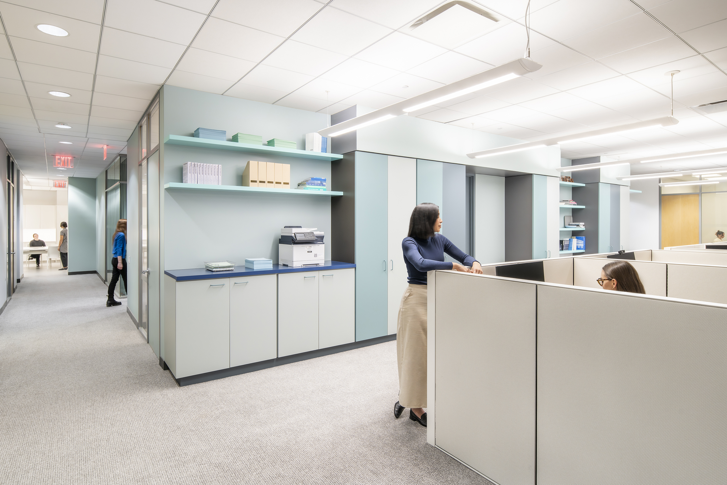 A view of the millwork cube with an open-plan cubicle workspace before it. In the foreground, a woman in a navy blue top and beige skirt leans on a cubicle partition, engaging in conversation with a seated colleague wearing glasses. To the left, a hallway leads to a glass-enclosed meeting room where two people are seated at a table. Another woman in a blue blouse walks past the meeting room. The office features a neutral color palette with light blue and gray tones, sleek cabinetry, open shelving with office supplies, and overhead lighting.