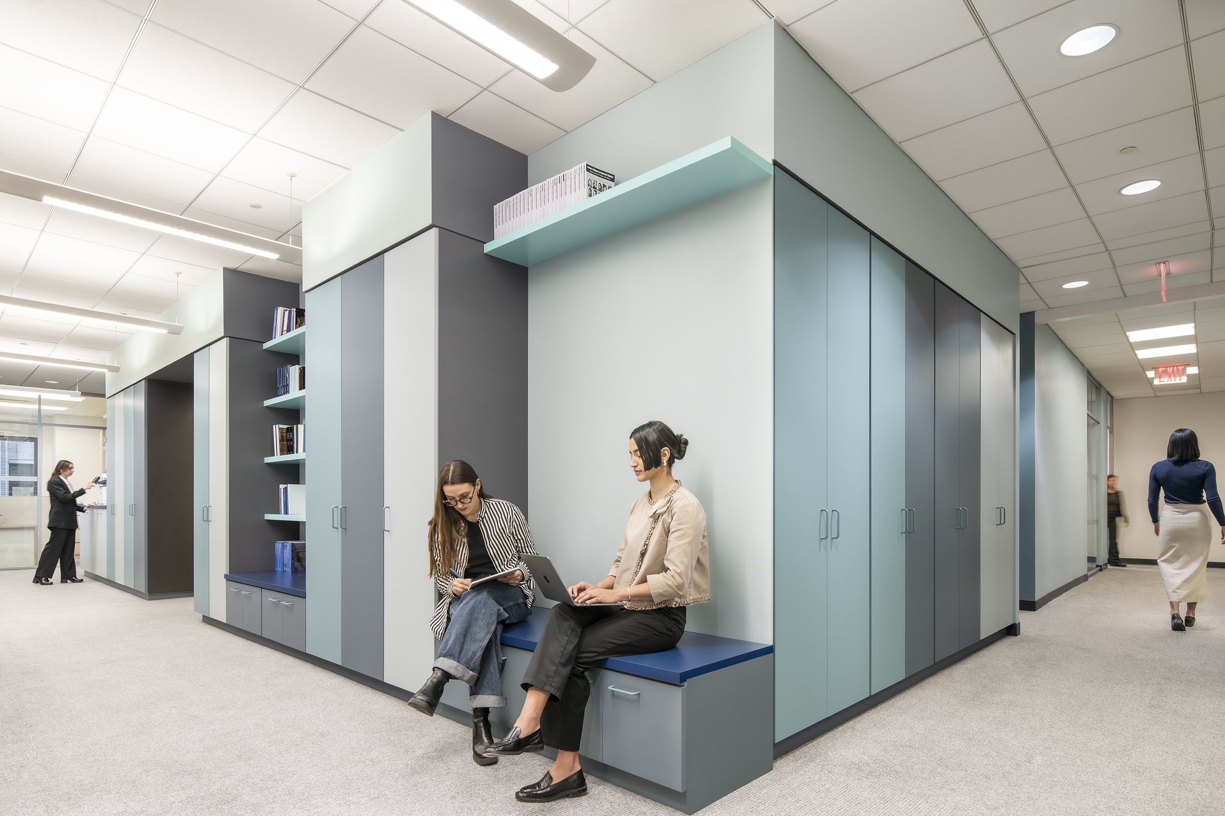 View of the central millwork cube with integrated bench seating in the foreground at the corner, a person making copies at the copy counter in the left background, and a person walking down the hallway to another person standing at the rear right background