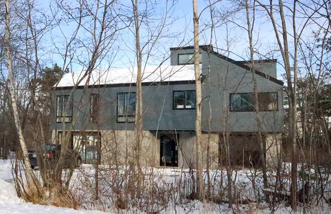 A modern house with a sloped roof and large windows, partially hidden by leafless trees in a snowy landscape.
