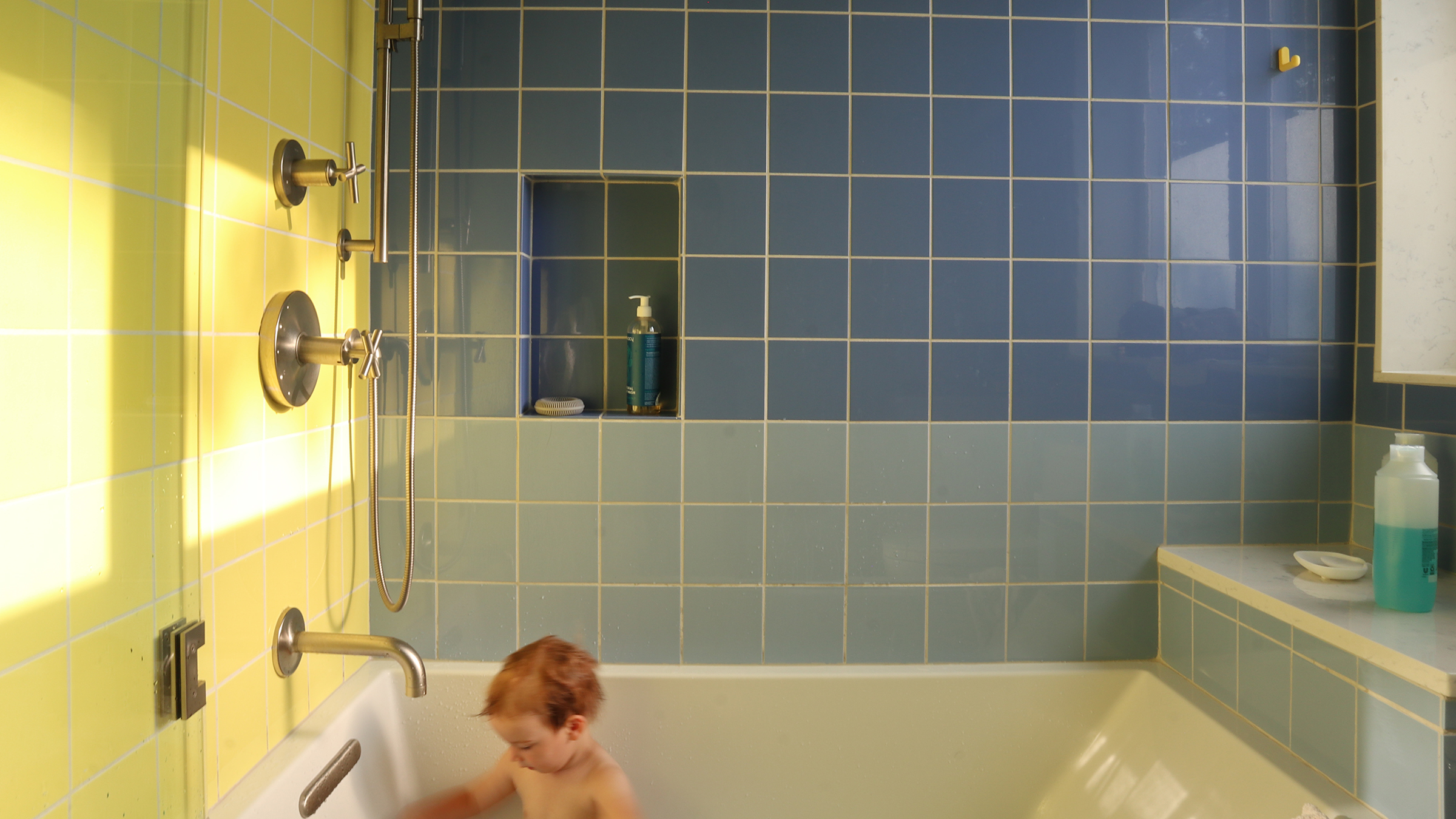 A bathroom with blue tiled wall and yellow tiled wall, featuring a geometric bathtub with contemporary showerhead and faucet fixtures. Sunlight casts a warm glow on the tiles. A young child with wet hair is sitting in the bathtub, playing with the water. Shelves in the tiled wall hold bottles of soap and shampoo. A countertop with additional toiletries is visible on the right side of the image.