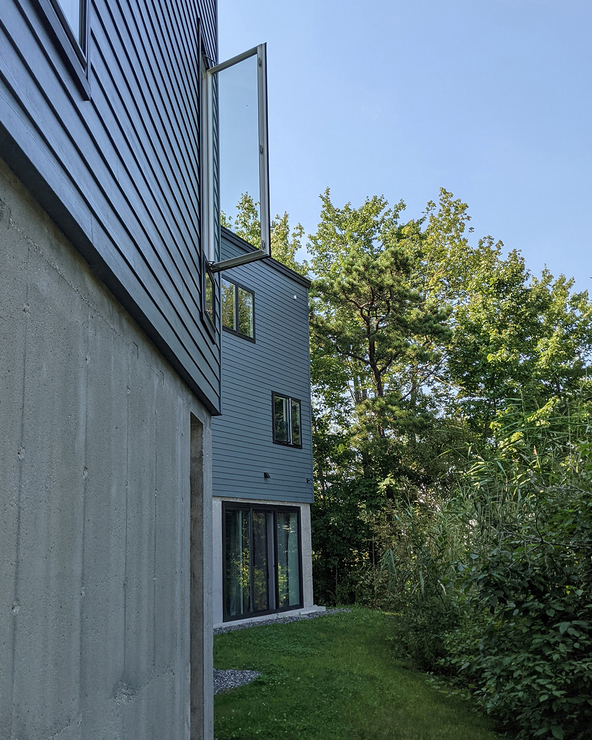 A house with an angular design, featuring cool blue horizontal siding, concrete walls, and an open glass window. Lush greenery stands in contrast to the clean vertical lines of the house