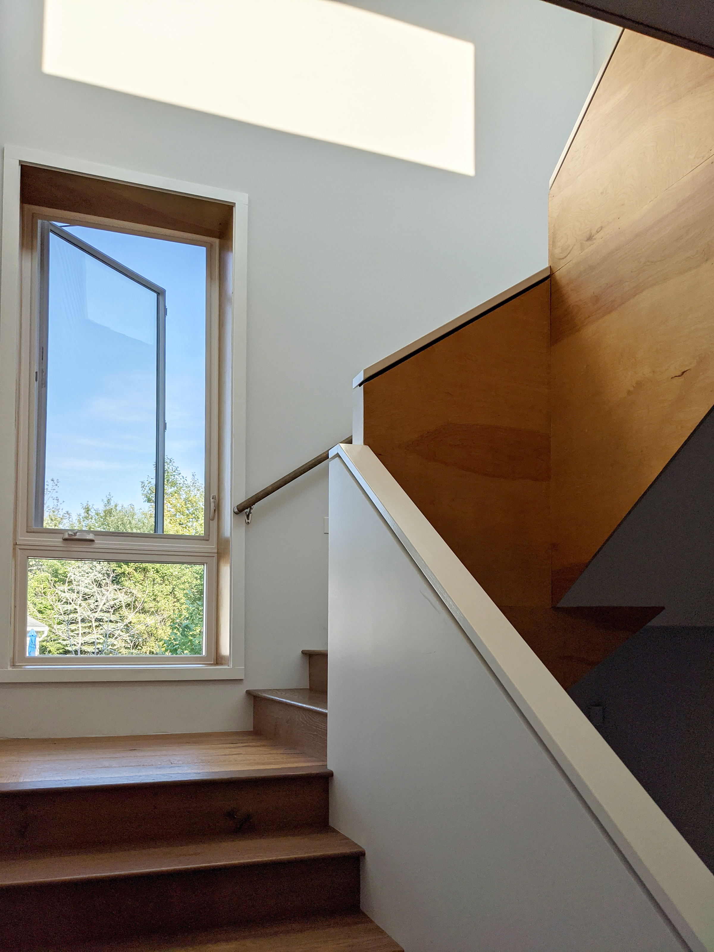 A wooden staircase with white railings leads upward, lit by sunlight from a large open window. A geometric shadow is cast on the white wall, and greenery is visible through the window.