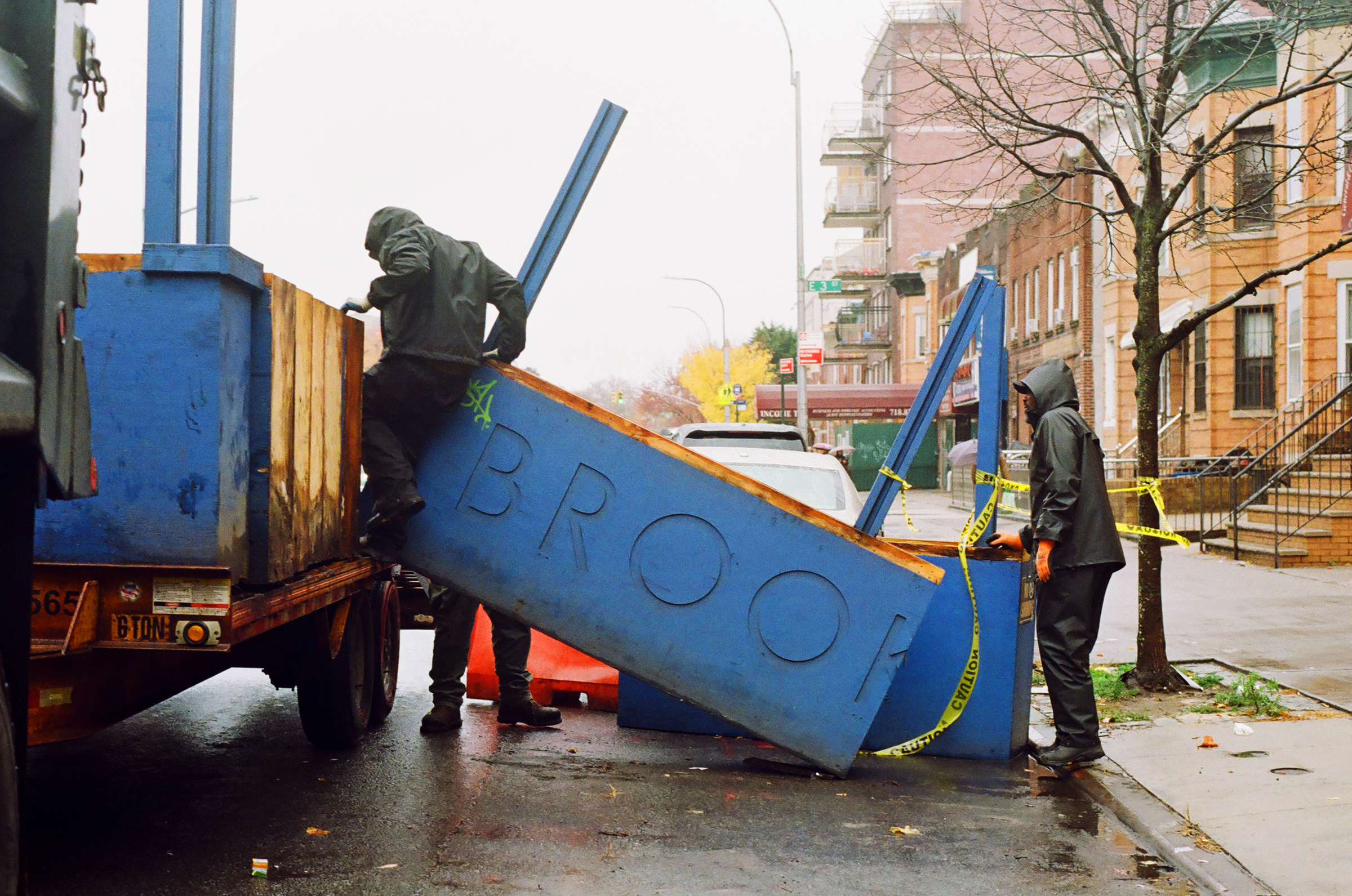 Two workers in rain gear loading a large section of a blue strucgture with the word "BROOK" from a wet city street onto a truck.