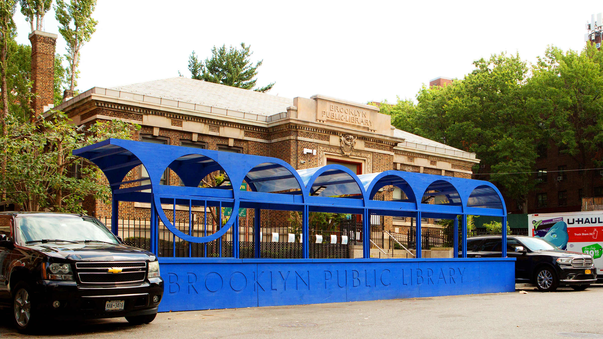 Photograph of the Brooklyn Public Library building with a distinctive blue wood and fabric structure, cars, and trees visible.