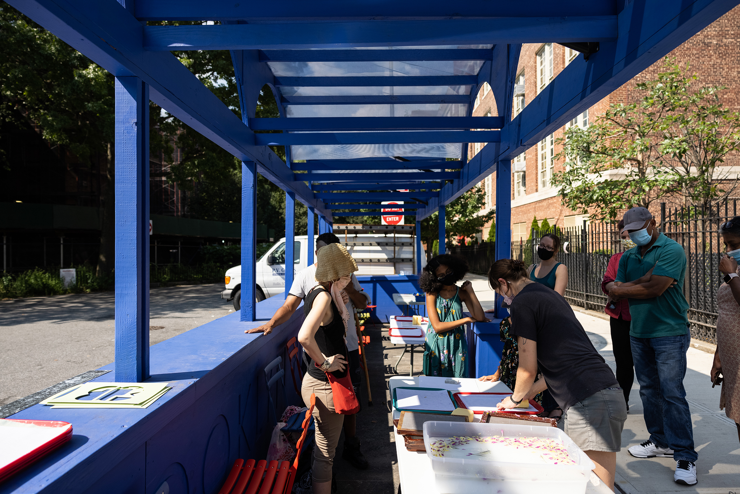 Group of people interacting at tables under a blue structure with a translucent roof, on a sunny day.