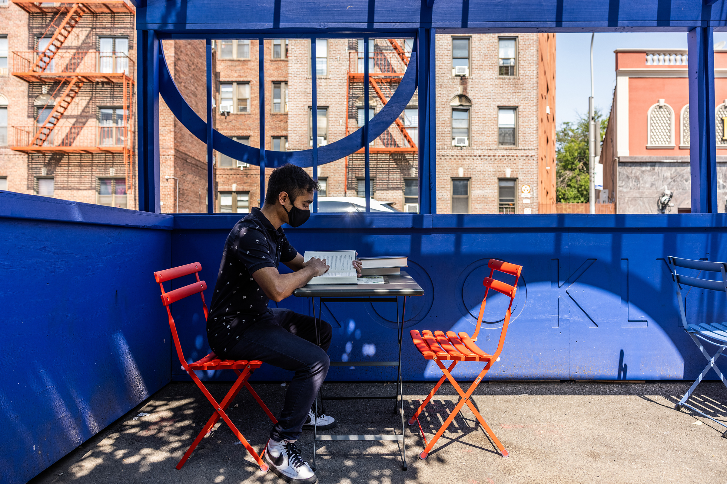 Individual wearing a mask seated at a table with books under a bright blue structure, with buildings in the background.