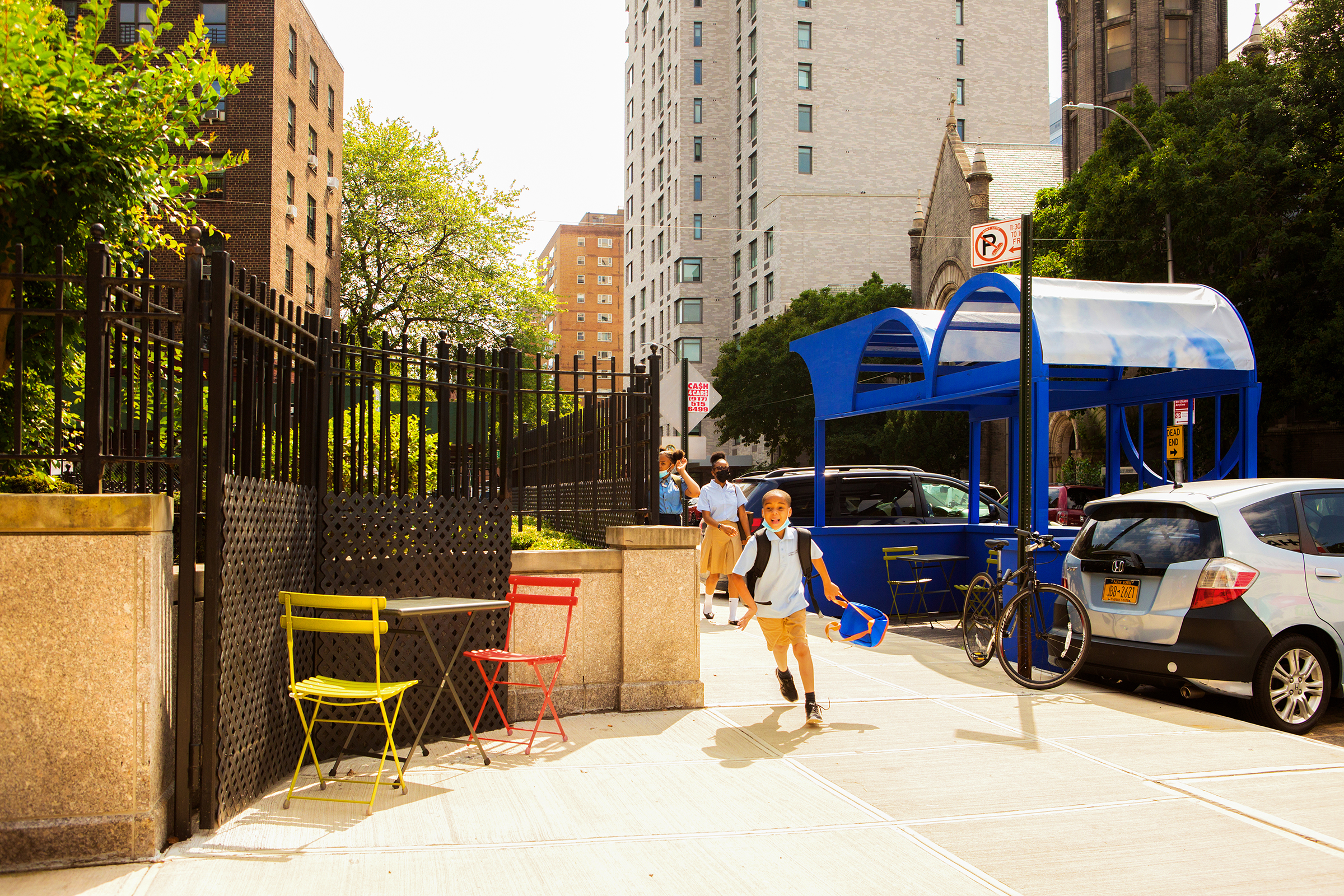 Young boy with a backpack running on a sunny sidewalk next to a black fence, with a blue public structure and parked cars in the background.