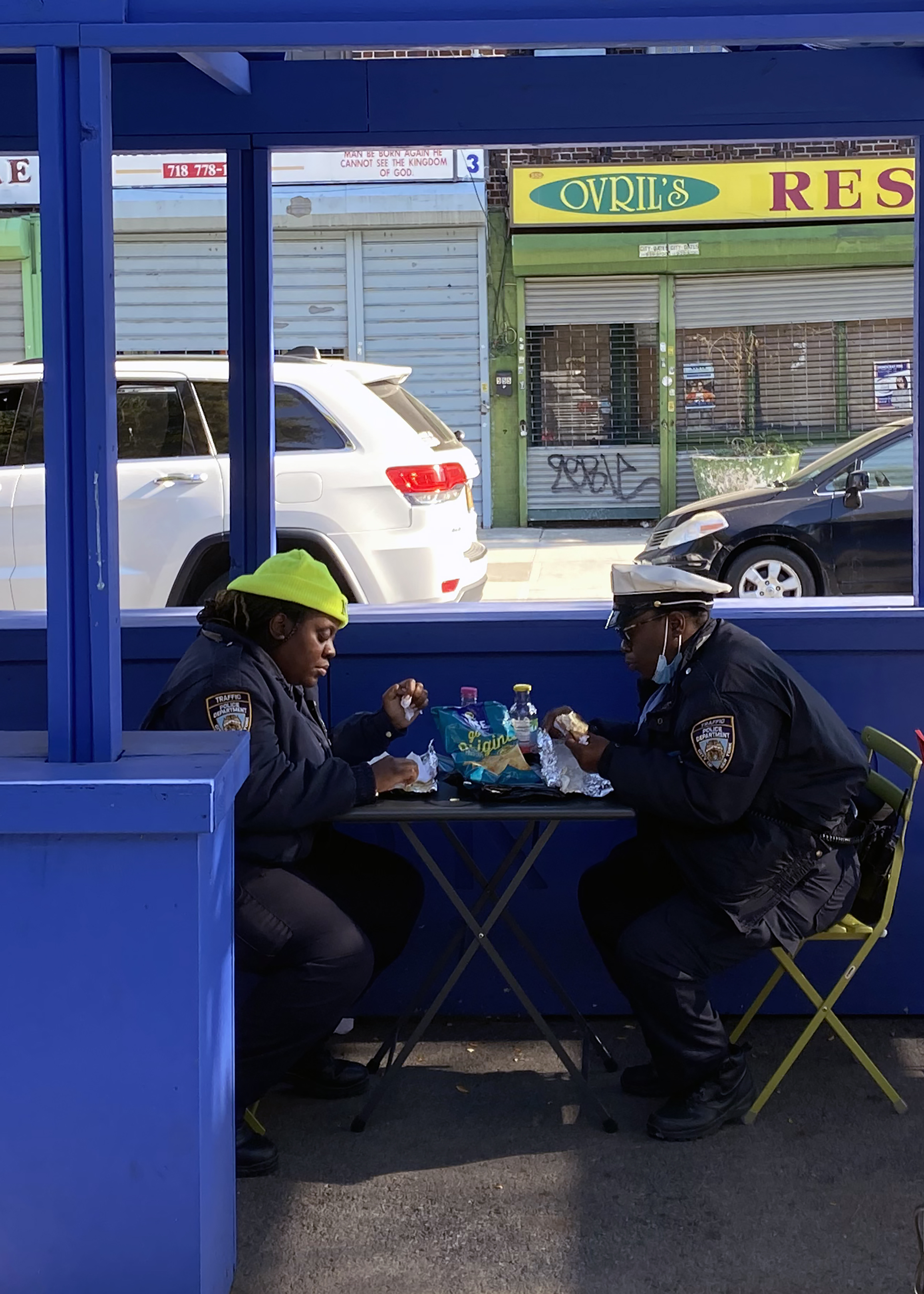 Two people in uniforms having lunch at a small table under a blue structure, with a street and storefronts in the background.